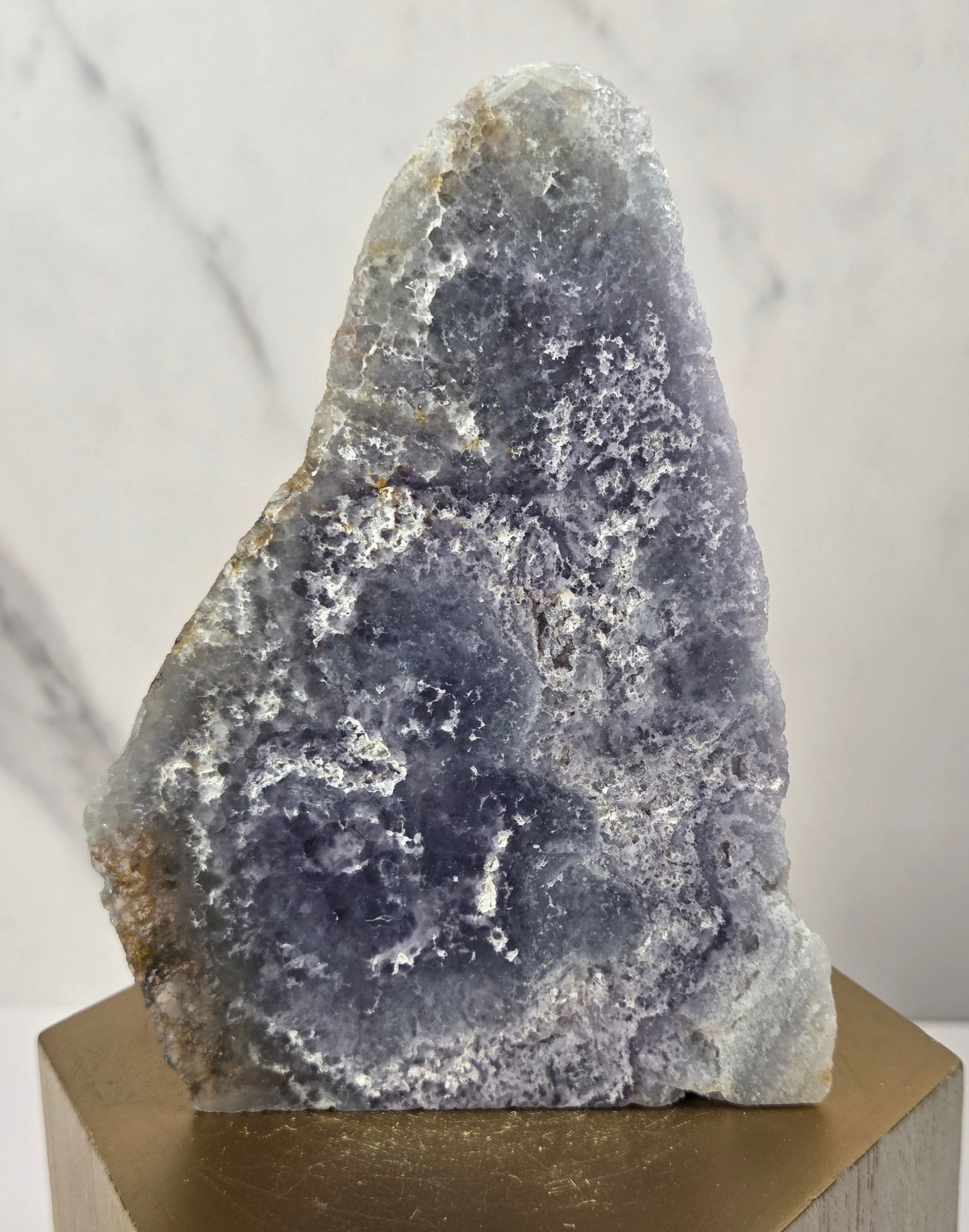 raw mineral and geode specimens displayed on wooden blocks against a white marble-patterned background. The stones feature natural patterns in shades of green, purple, blue, and gray.