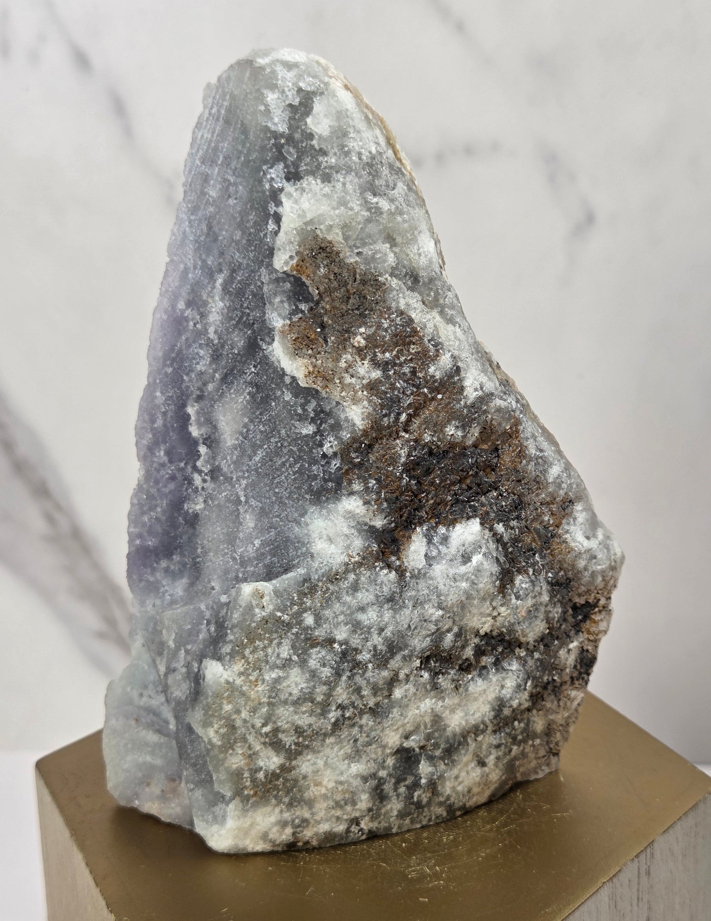 raw mineral and geode specimens displayed on wooden blocks against a white marble-patterned background. The stones feature natural patterns in shades of green, purple, blue, and gray.