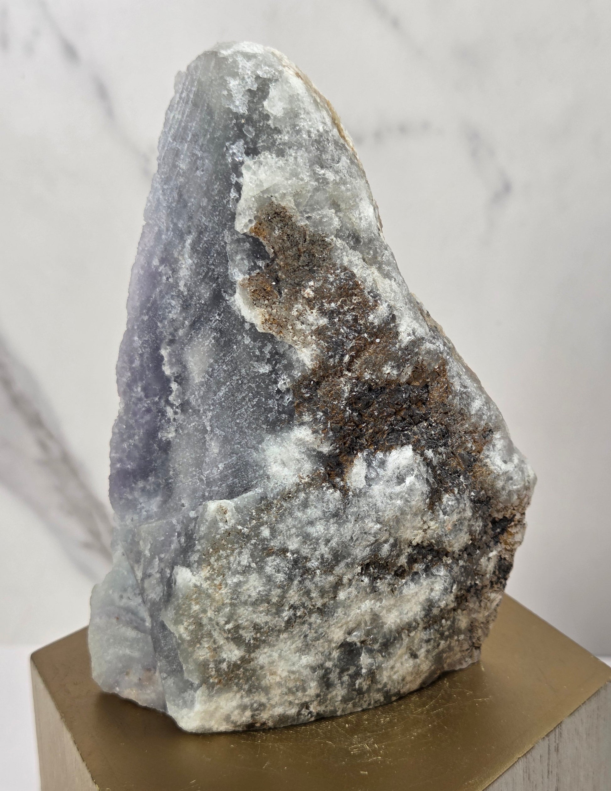 raw mineral and geode specimens displayed on wooden blocks against a white marble-patterned background. The stones feature natural patterns in shades of green, purple, blue, and gray.