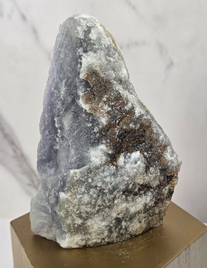 raw mineral and geode specimens displayed on wooden blocks against a white marble-patterned background. The stones feature natural patterns in shades of green, purple, blue, and gray.