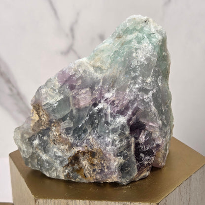 raw mineral and geode specimens displayed on wooden blocks against a white marble-patterned background. The stones feature natural patterns in shades of green, purple, blue, and gray.