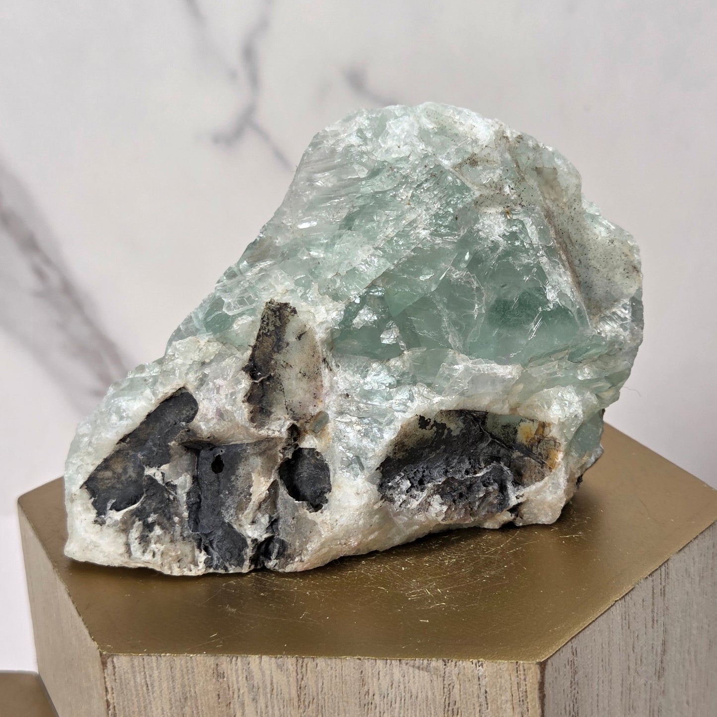 A raw mineral specimen with pale green translucent crystal and dark black inclusions, displayed on a gold-colored geometric pedestal against a light marble-patterned background.