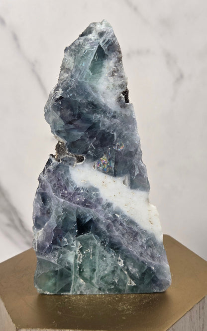 A raw fluorite crystal specimen with blue, green, and purple bands, displayed on a gold hexagonal pedestal against a light marble background.