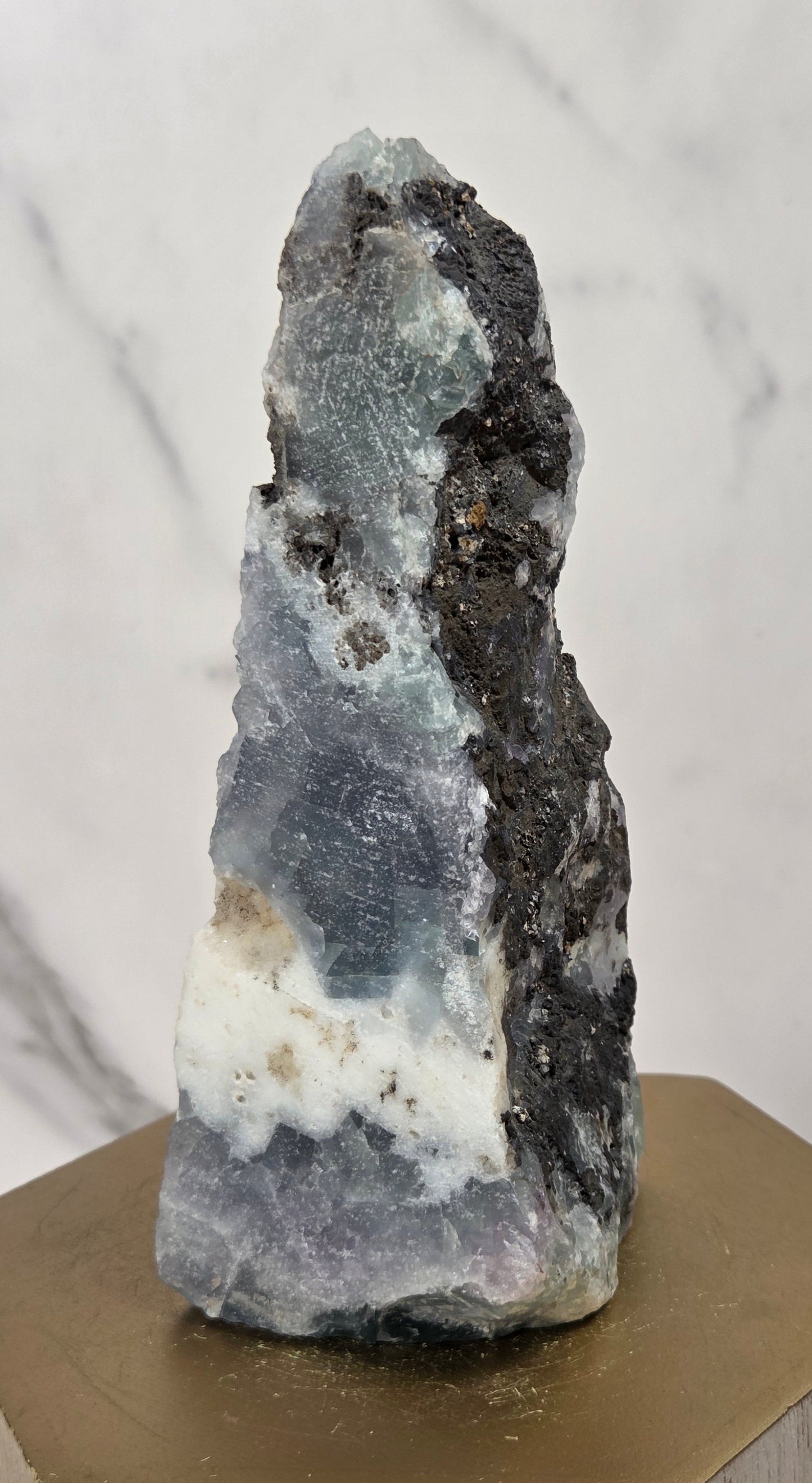 A natural mineral specimen with layered blue-gray, white, and dark charcoal sections, displayed on a hexagonal gold-toned wooden block against a light marble-patterned background.