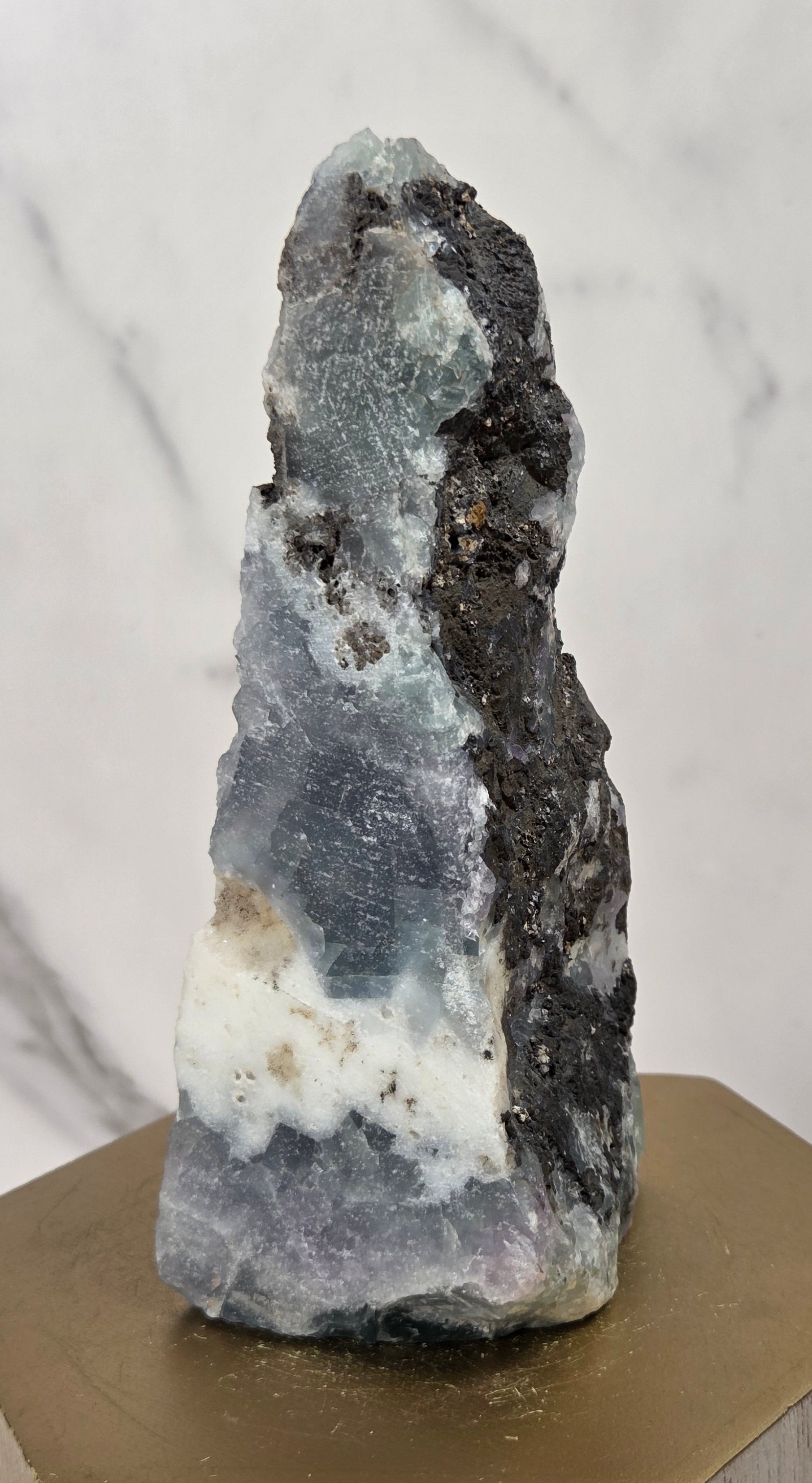 A natural mineral specimen with layered blue-gray, white, and dark charcoal sections, displayed on a hexagonal gold-toned wooden block against a light marble-patterned background.
