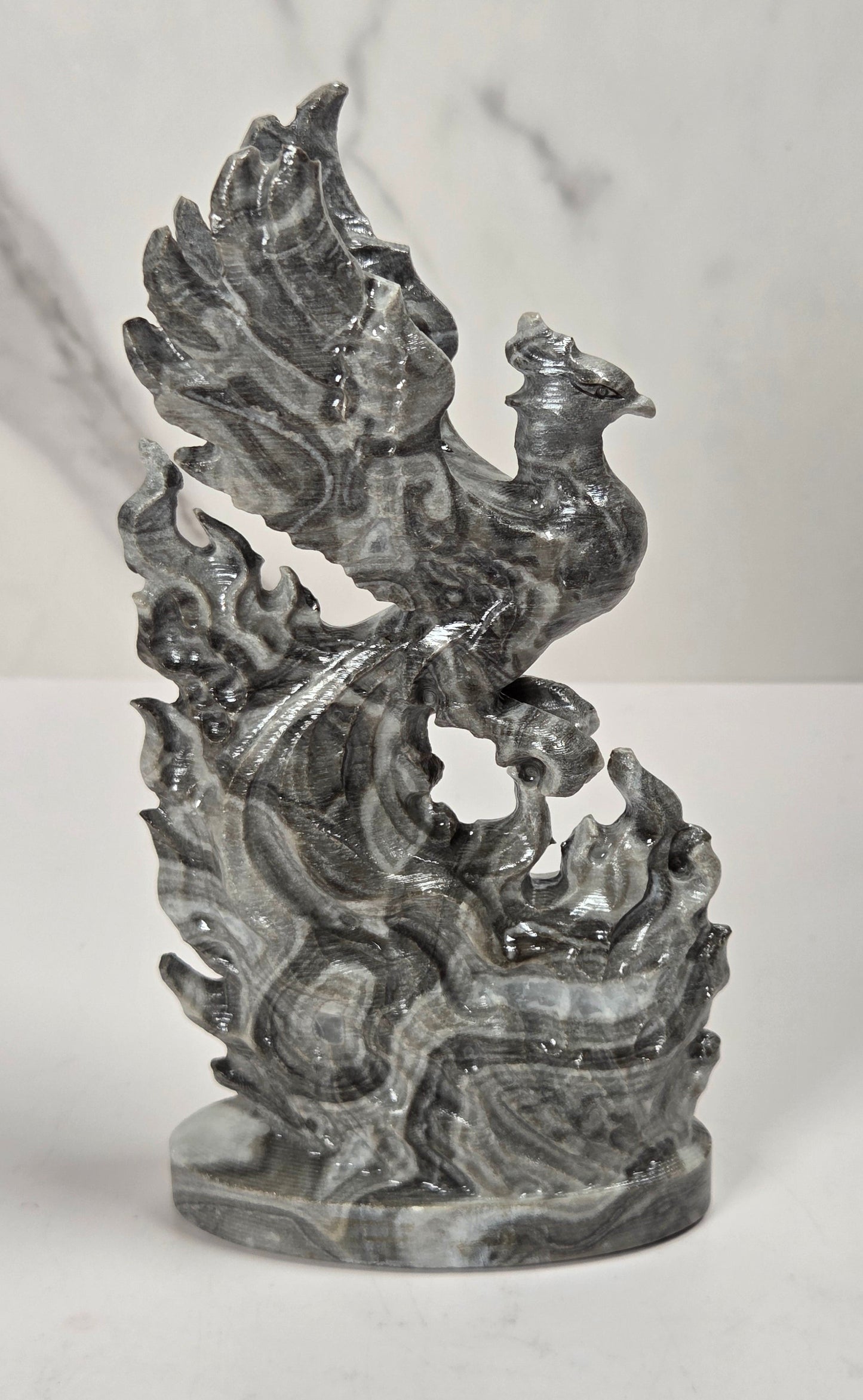 A dark gray stone phoenix sculpture standing on a circular base, photographed against a light marble-patterned background.