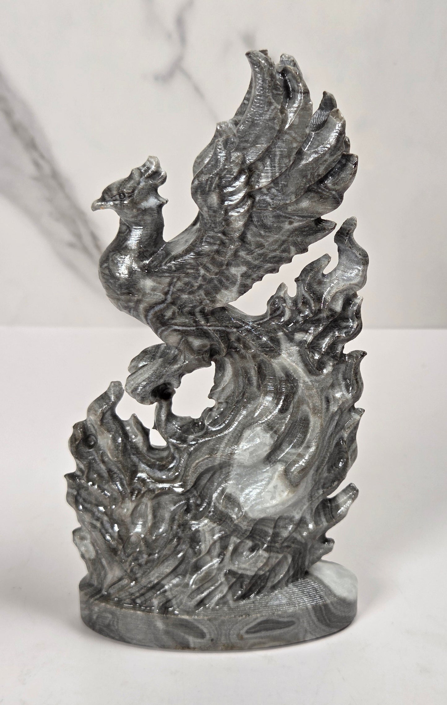 A metallic silver sculpture of a phoenix rising from stylized flames, displayed on a white marble surface against a white marble background.