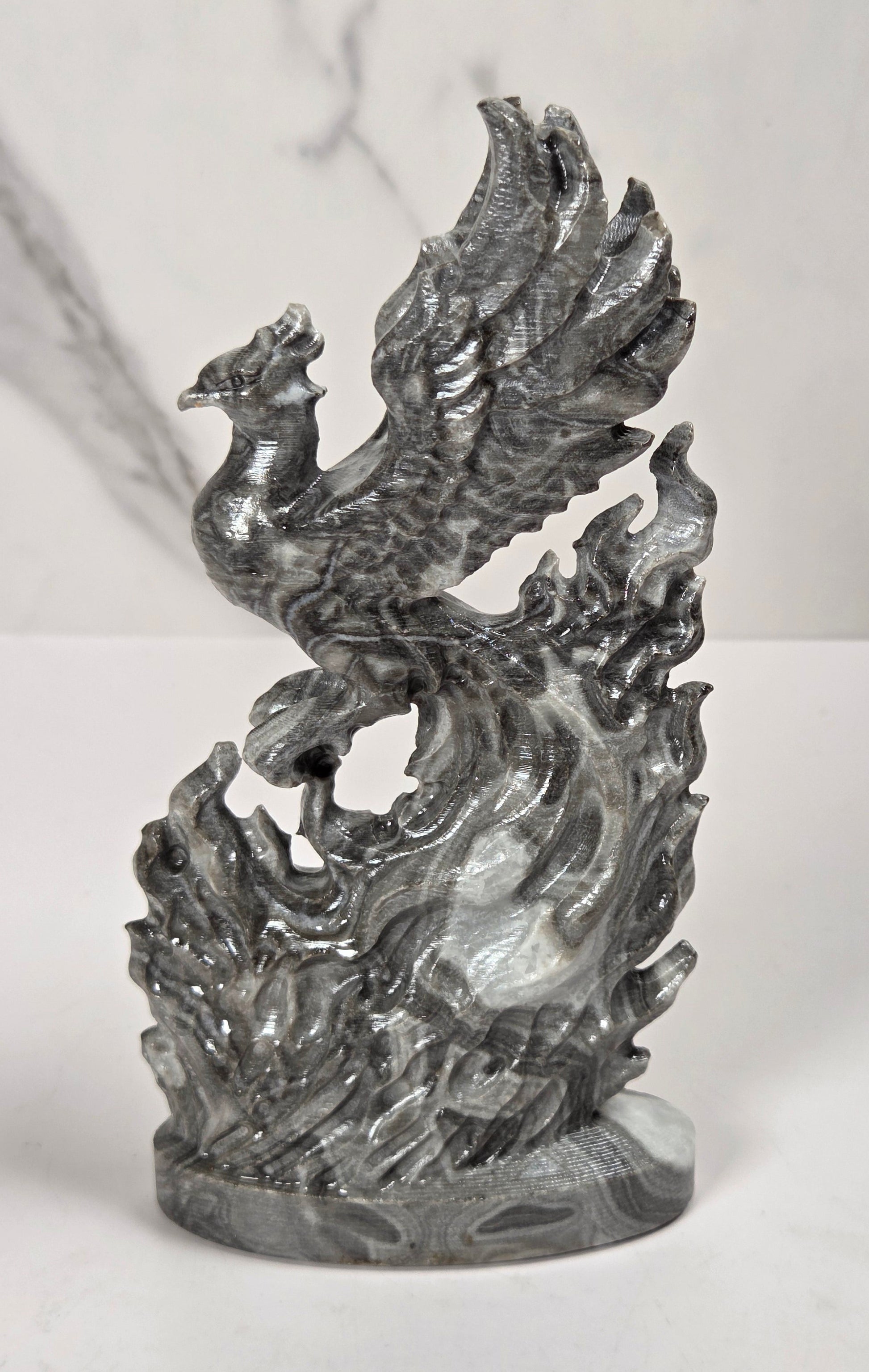 A metallic silver sculpture of a phoenix rising from stylized flames, displayed on a white marble surface against a white marble background.