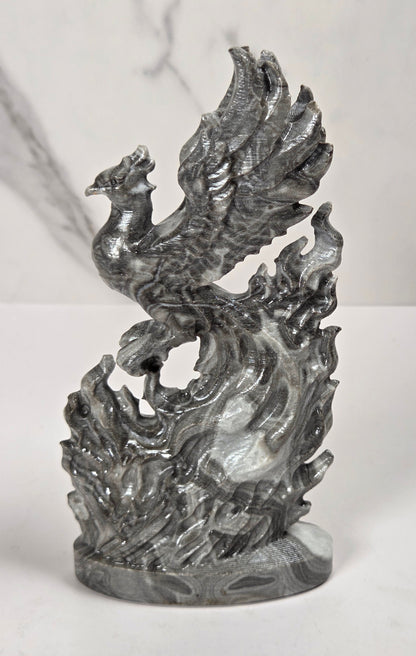 A metallic silver sculpture of a phoenix rising from stylized flames, displayed on a white marble surface against a white marble background.