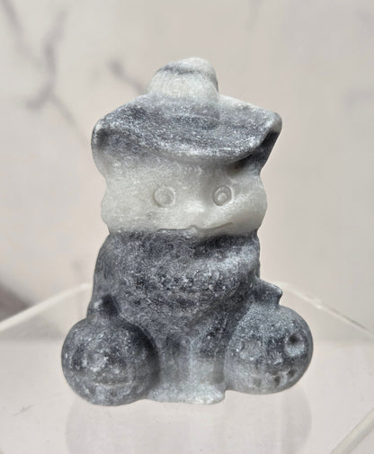 A small gray and white speckled figurine of a seated character wearing a hat, displayed on a clear acrylic stand against a light marble-patterned background.