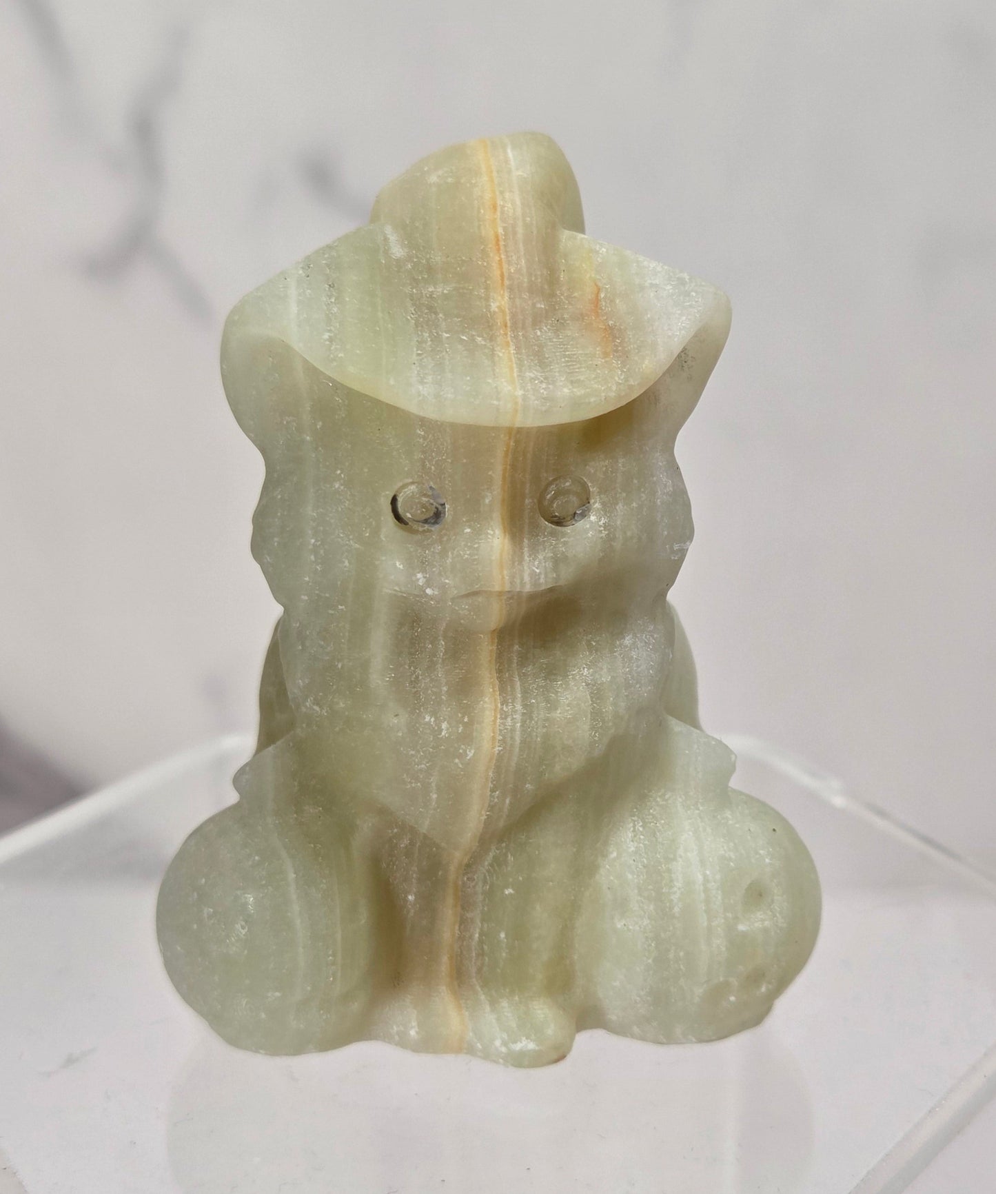 A small carved figurine of a seated animal wearing a hat, made from pale translucent stone with subtle yellow and green tones, displayed on a clear acrylic stand against a light marble-patterned background.