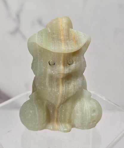 A small carved figurine of a seated animal wearing a hat, made from pale translucent stone with subtle yellow and green tones, displayed on a clear acrylic stand against a light marble-patterned background.