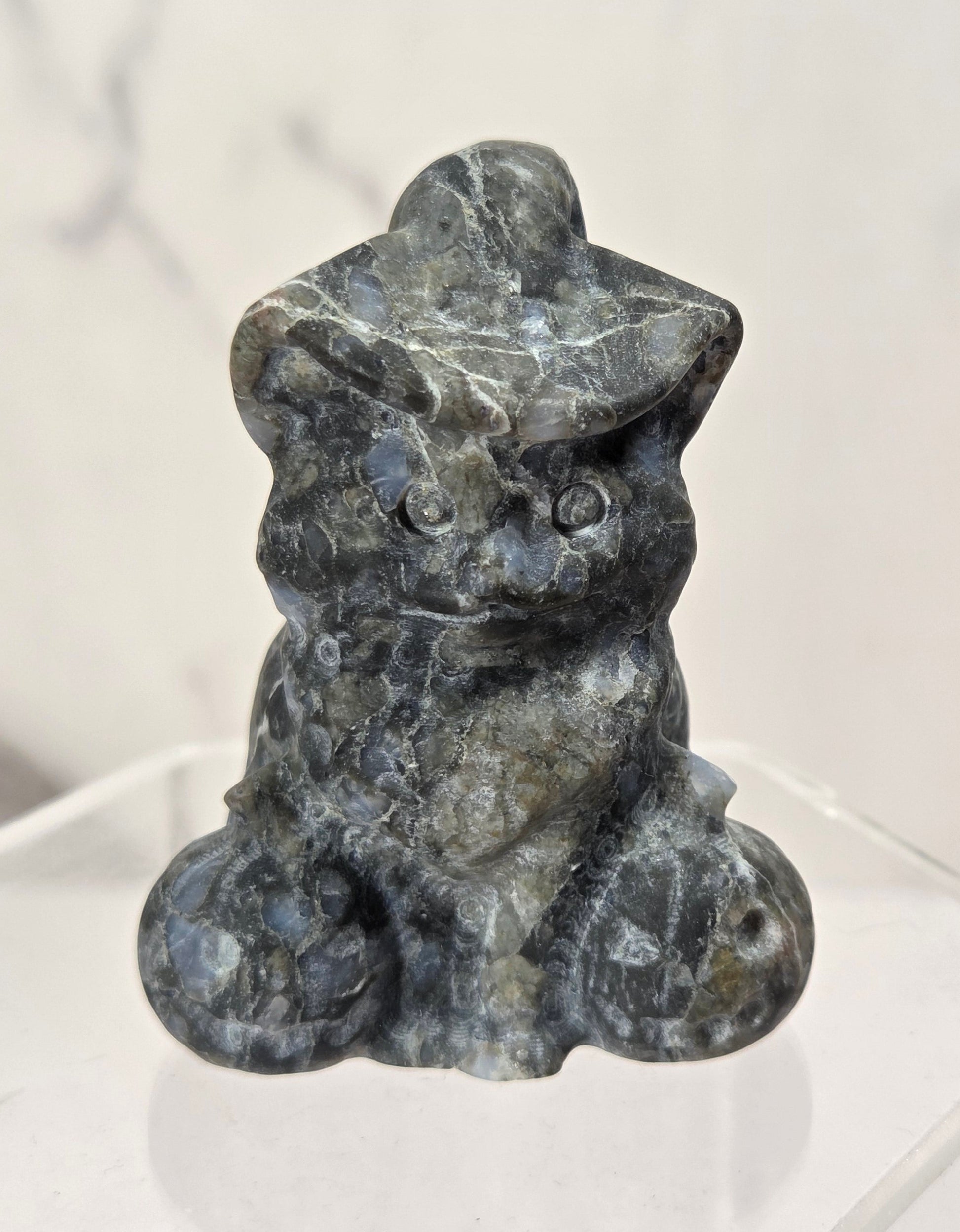 A small carved stone figurine of a seated cat wearing a hat, shown on a clear acrylic display base against a light marble-patterned background.