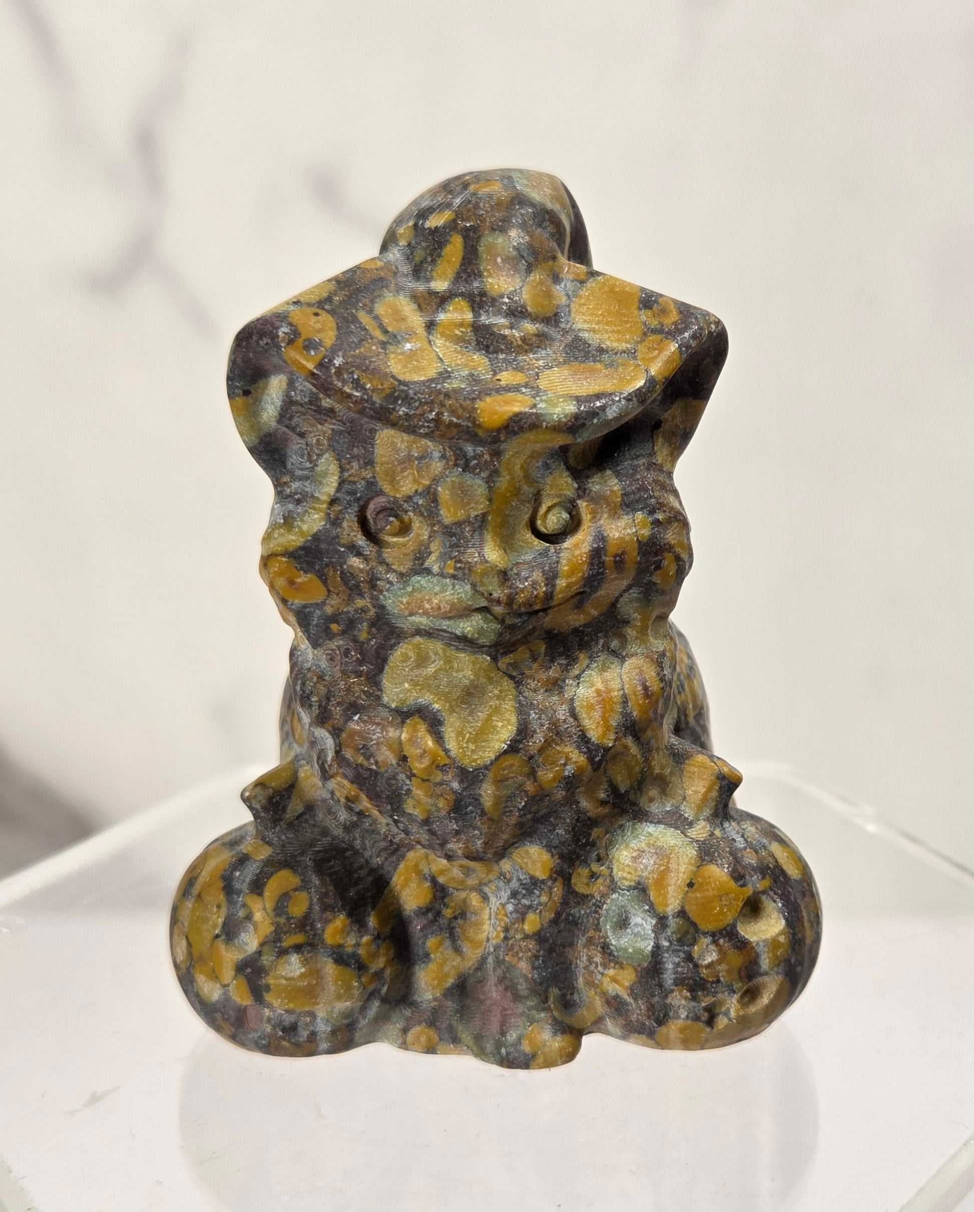 A small carved figurine of a seated cat wearing a hat, made from mottled stone with dark gray, black, and golden yellow patterns, displayed on a clear acrylic hexagonal pedestal against a light gray marble-patterned background.