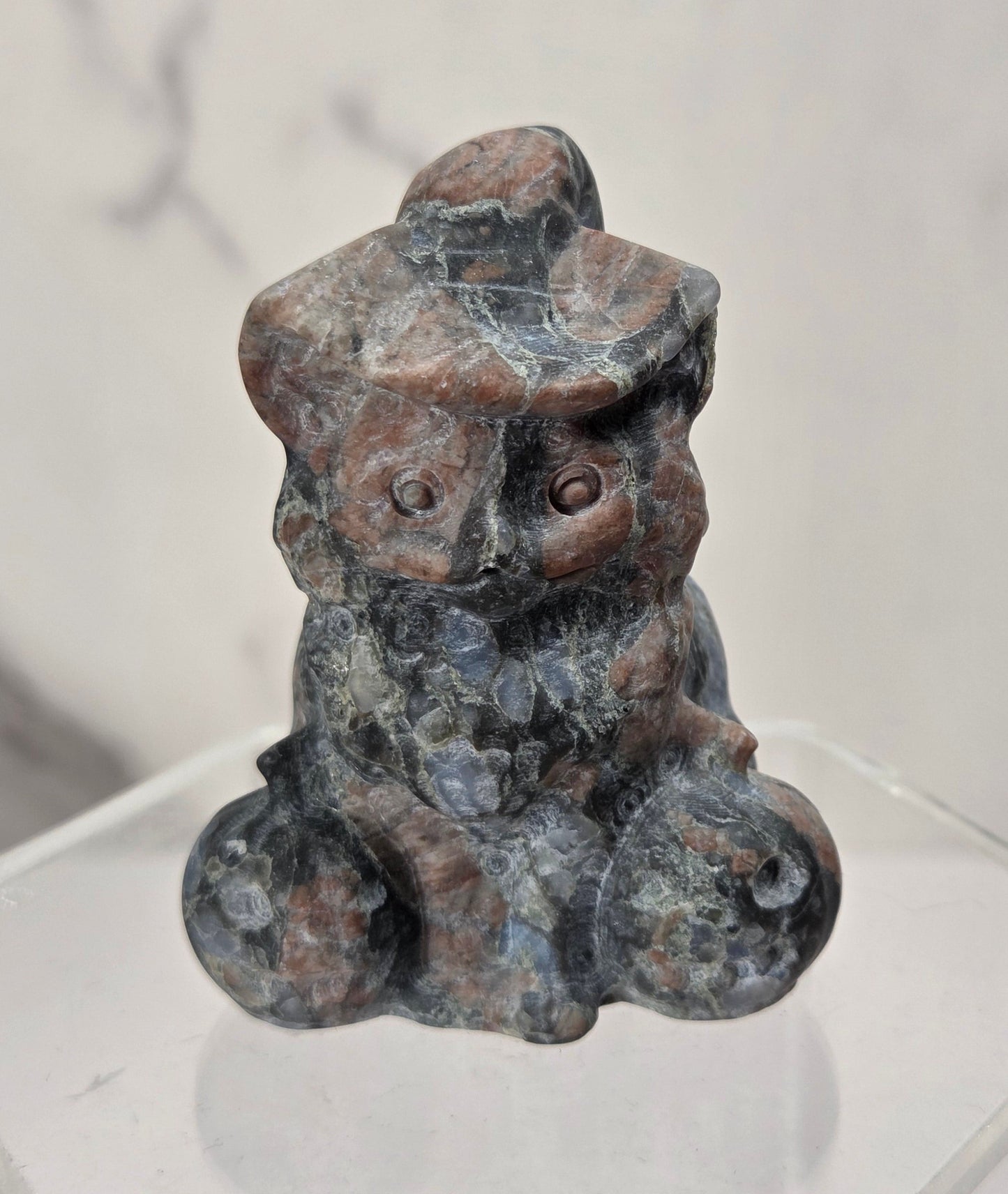 A small carved stone figurine of a seated figure with a hat, shown on a clear acrylic display base against a light marble-patterned background.