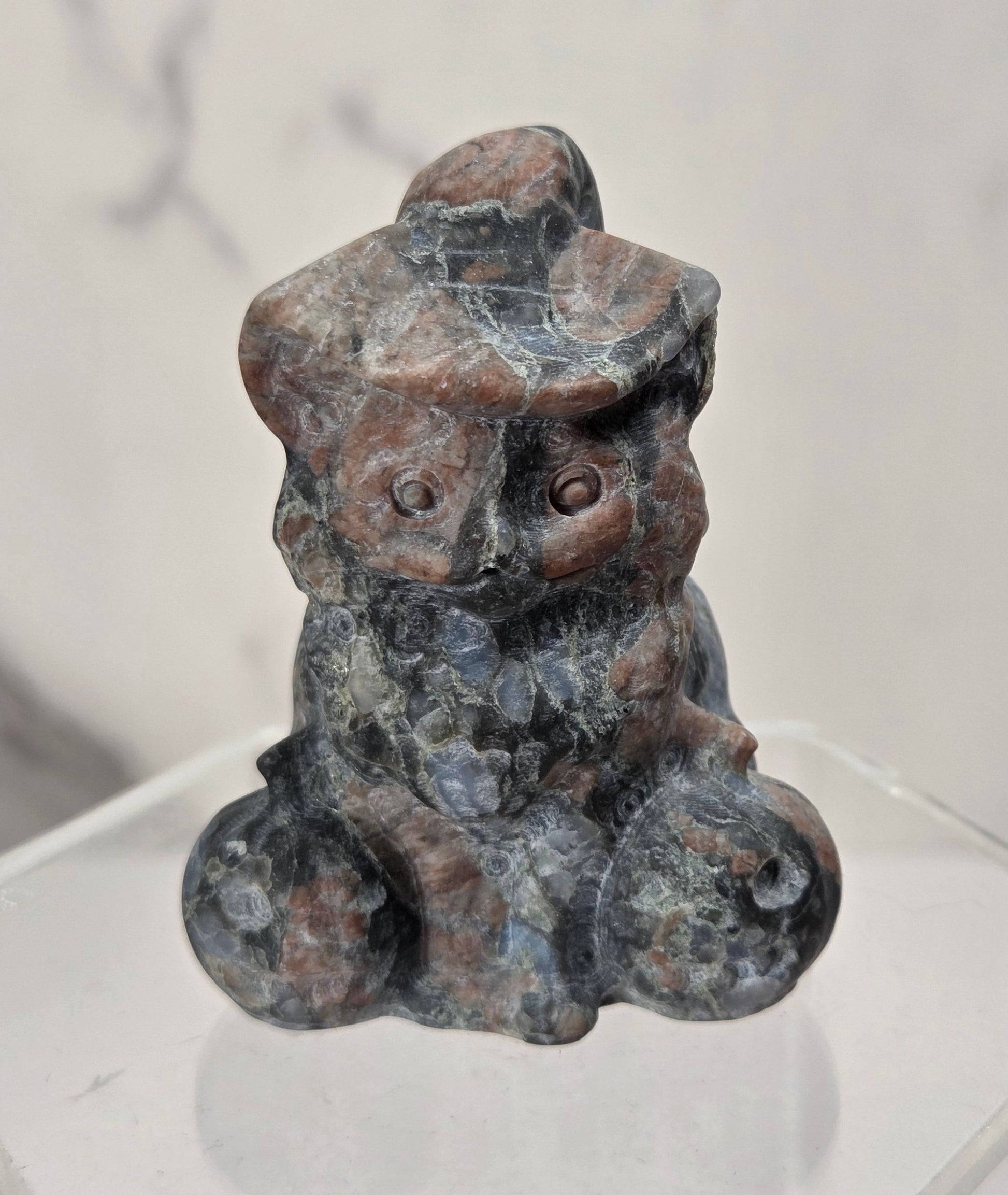 A small carved stone figurine of a seated figure with a hat, shown on a clear acrylic display base against a light marble-patterned background.