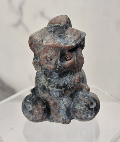 A small carved stone figurine of a seated figure with a hat, shown on a clear acrylic display base against a light marble-patterned background.