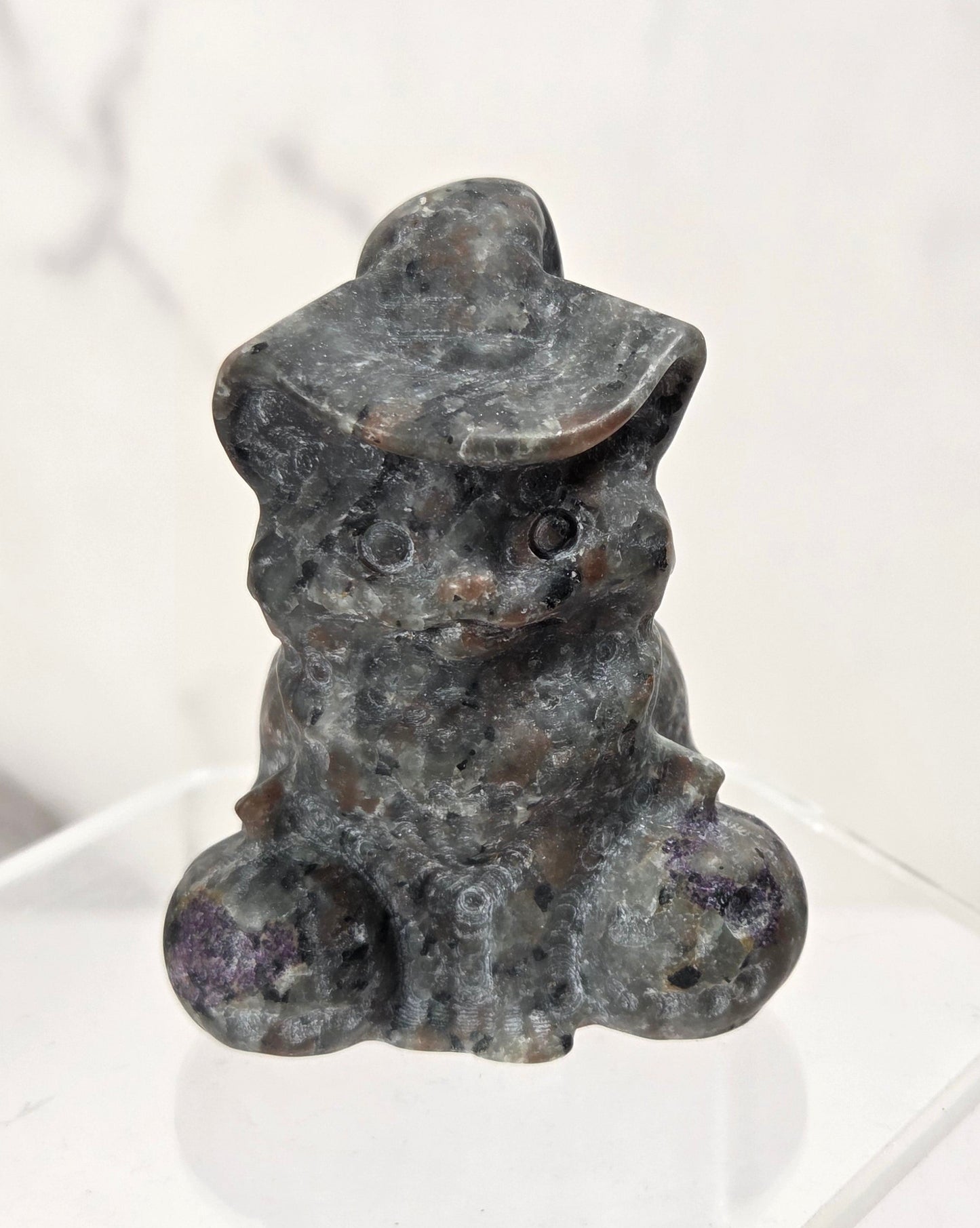A small carved stone figurine of a seated rabbit wearing a hat, displayed on a clear acrylic stand against a light marble-patterned background.