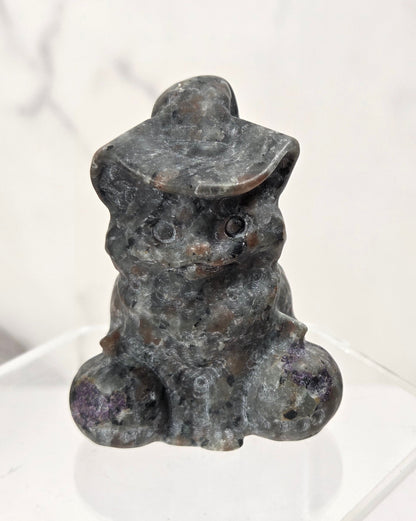 A small carved stone figurine of a seated rabbit wearing a hat, displayed on a clear acrylic stand against a light marble-patterned background.