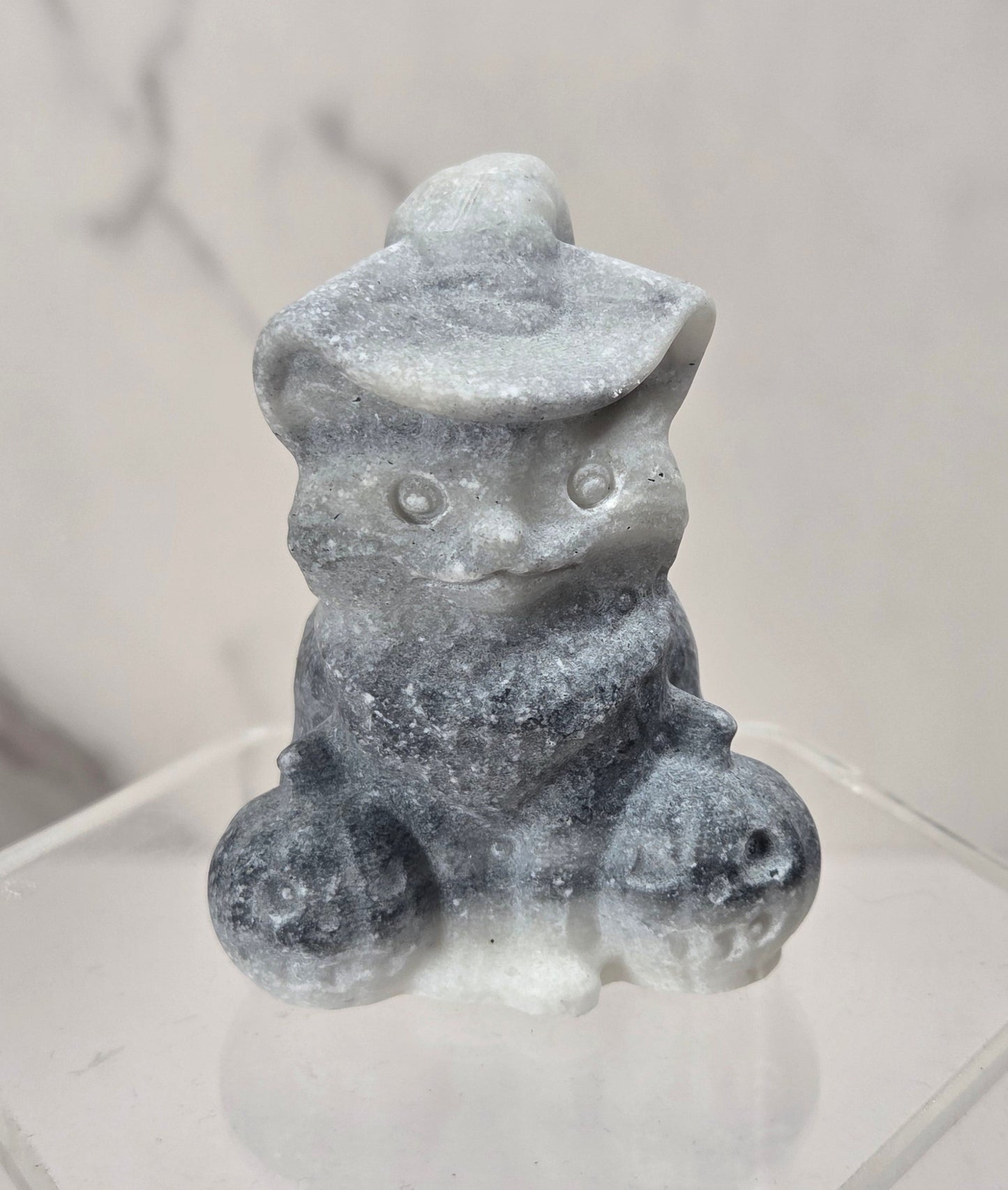 A small gray stone figurine of a seated cat wearing a hat, displayed on a clear acrylic hexagonal stand against a light marble-patterned background.