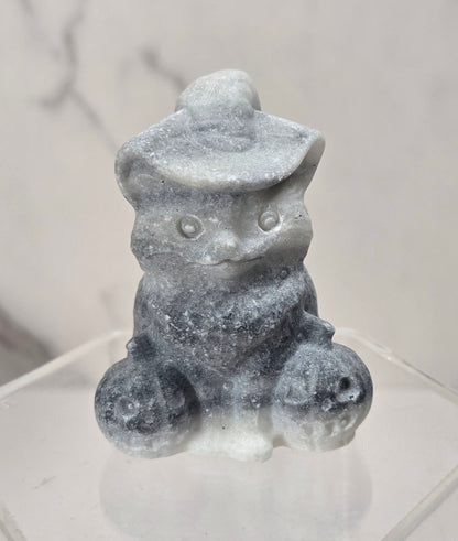 A small gray stone figurine of a seated cat wearing a hat, displayed on a clear acrylic hexagonal stand against a light marble-patterned background.