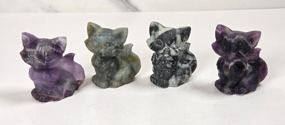 Four small carved stone animal figurines, including cats and a fox, displayed in shades of purple, gray, and black on a white marble surface.