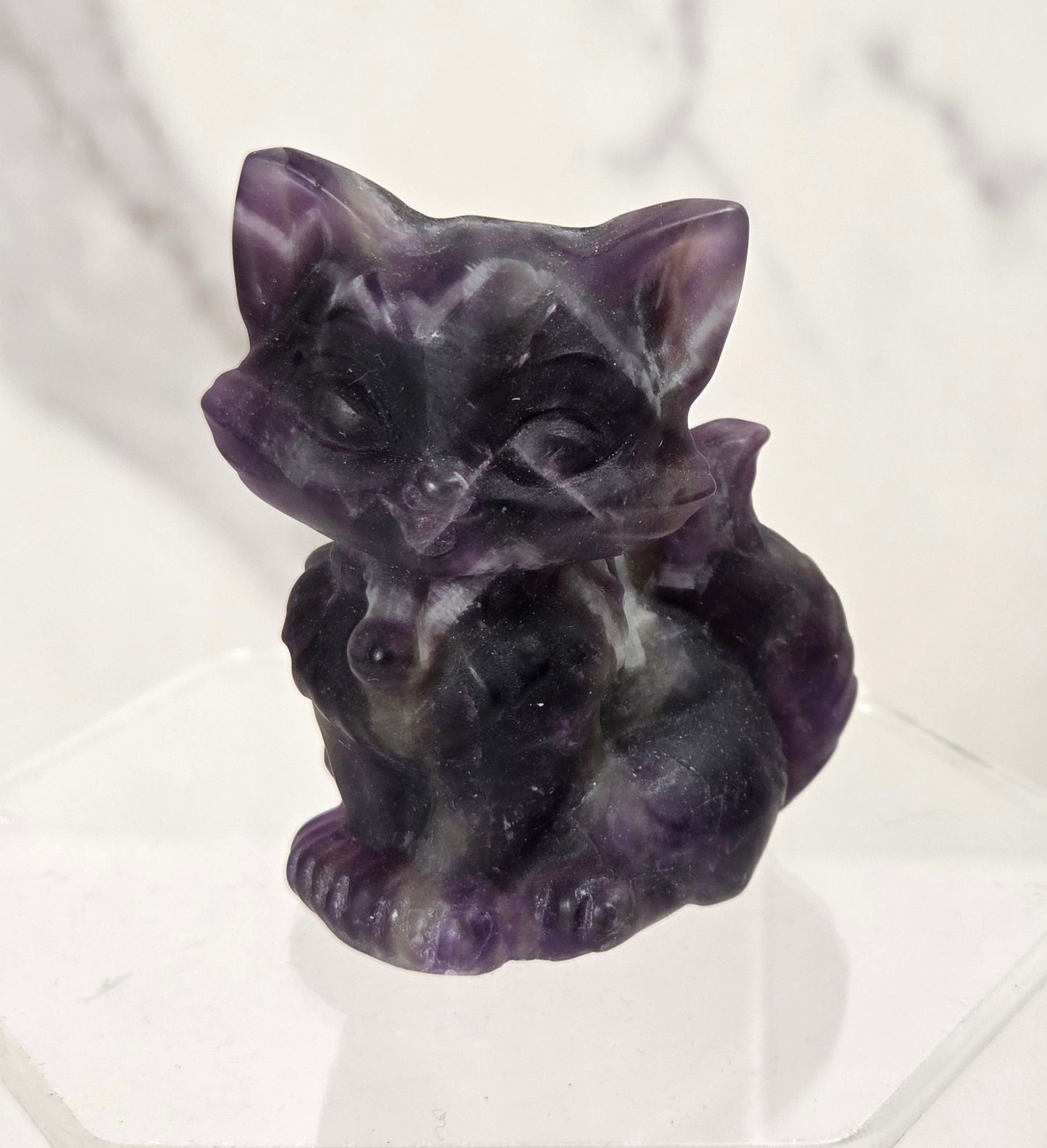 A small carved purple crystal cat figurine sitting on a clear acrylic display block against a white marble background.