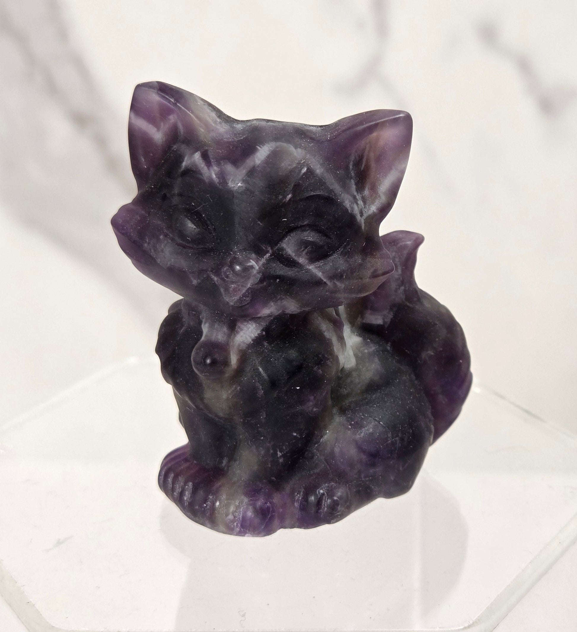 A small carved purple crystal cat figurine sitting on a clear acrylic display block against a white marble background.