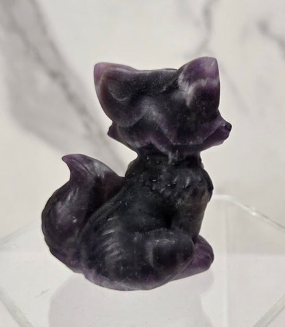 A small carved purple fox figurine sitting on a clear acrylic display block, set against a light gray marble-patterned background.