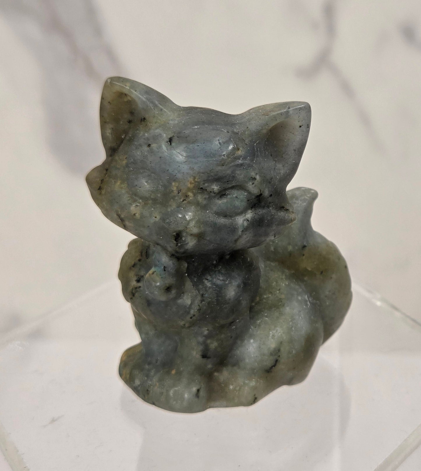 A small carved figurine of a cat in a crouched pose, made from a speckled gray-green stone, displayed on a clear acrylic hexagonal stand against a light marble-patterned background.