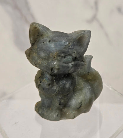 A small carved figurine of a cat in a crouched pose, made from a speckled gray-green stone, displayed on a clear acrylic hexagonal stand against a light marble-patterned background.
