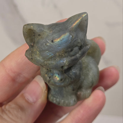 A small carved figurine of a cat made from iridescent labradorite stone, held in a person's hand against a light marble background.