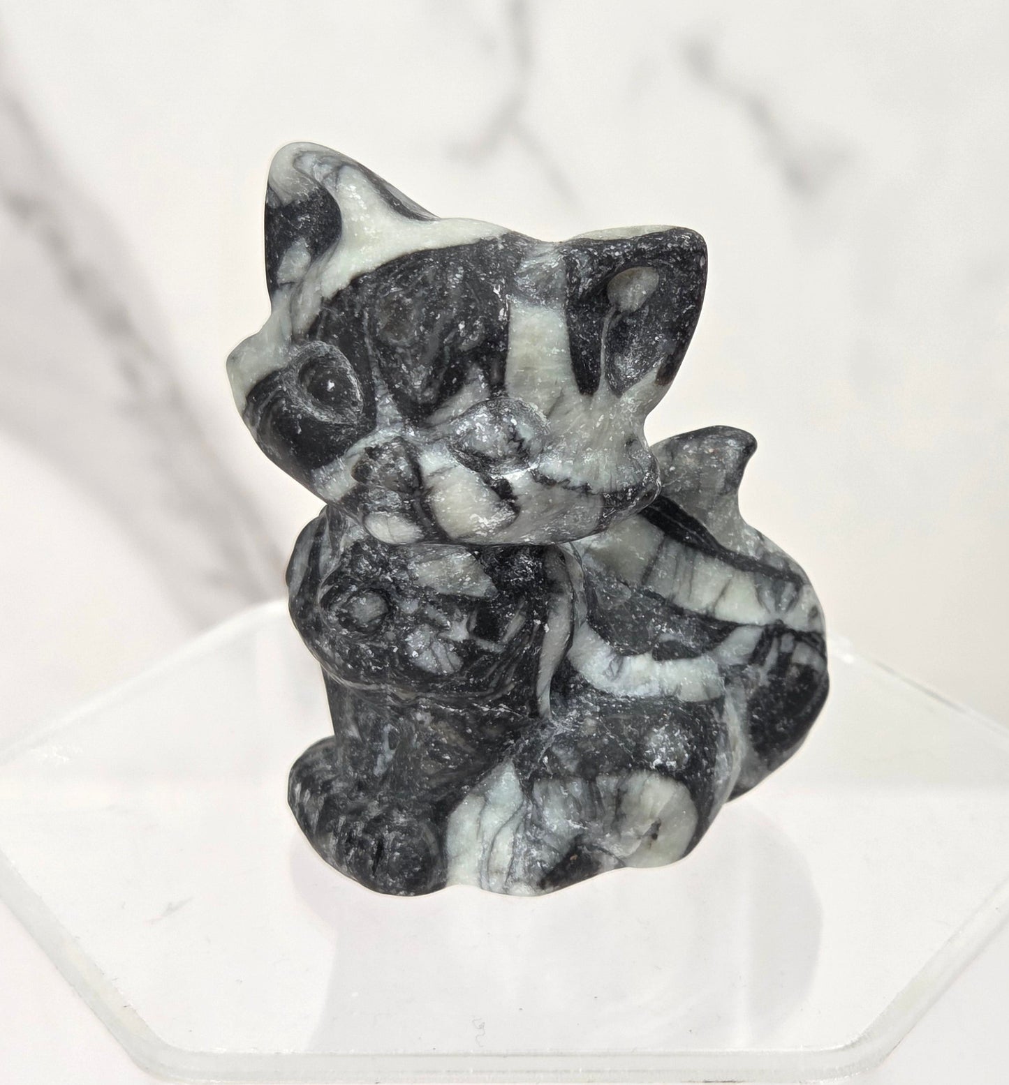 A small carved figurine of a cat holding a flower, made from black and white marbled stone, displayed on a clear acrylic stand against a white marble-patterned background.