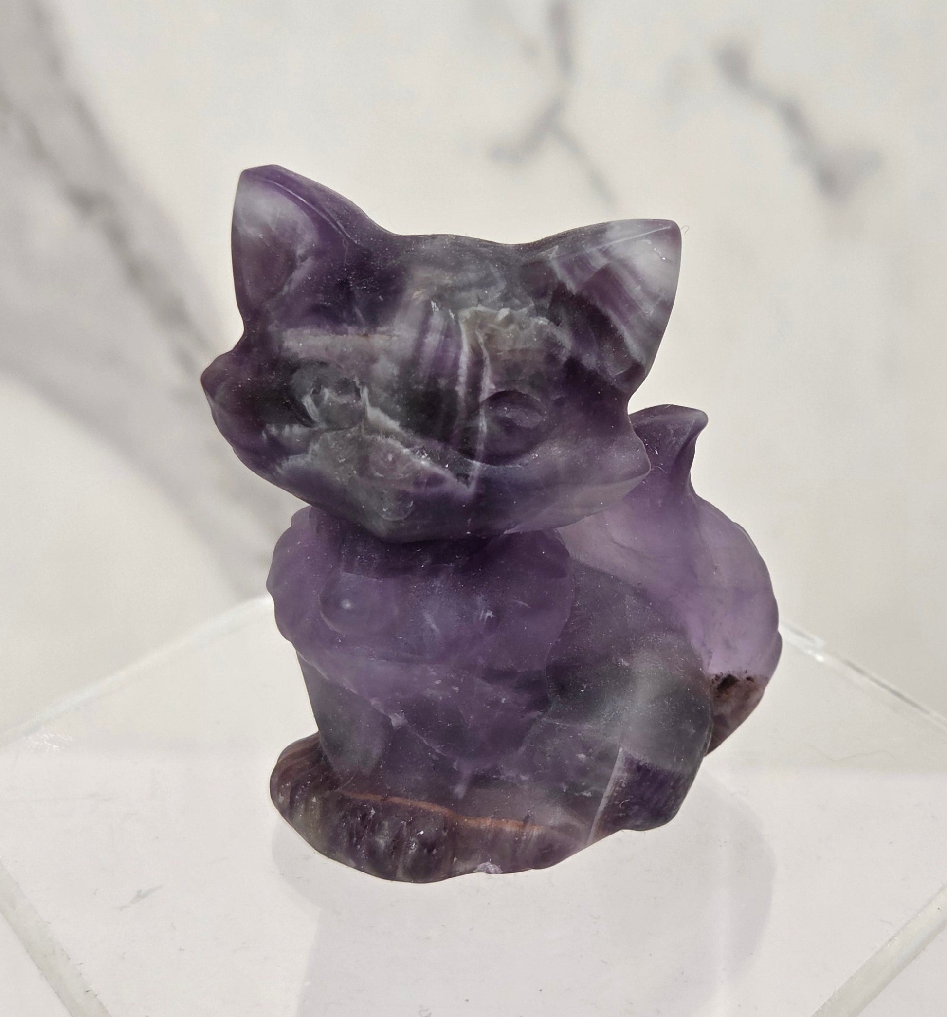 A small carved purple crystal cat figurine displayed on a clear acrylic stand against a light marble-patterned background.
