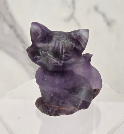 A small carved purple crystal cat figurine displayed on a clear acrylic stand against a light marble-patterned background.