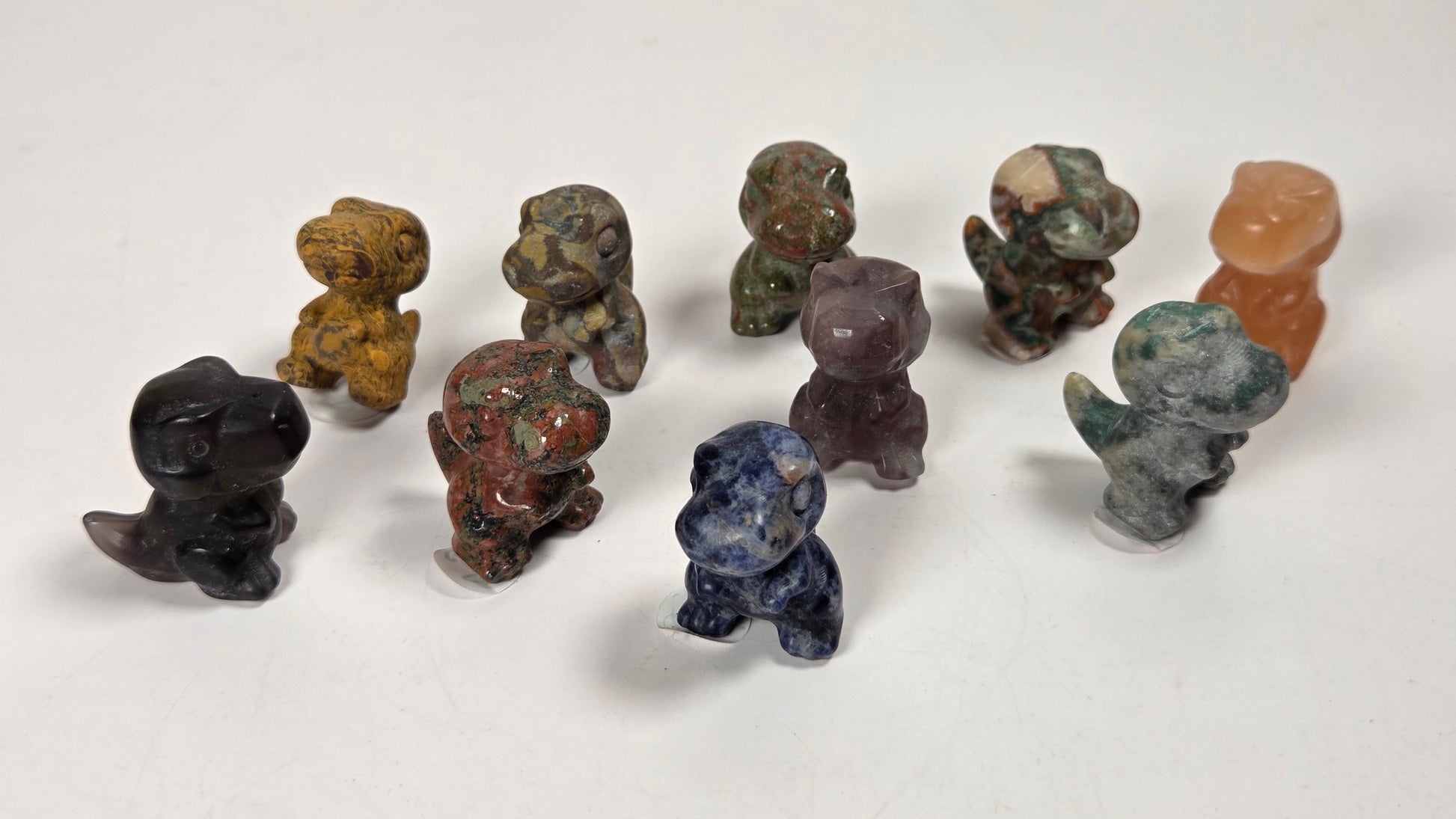 A group of small carved stone figurines shaped like dinosaurs, displayed on a plain white surface. The figurines are made from various natural stones in different colors, including black, brown, green, blue, purple, and orange.