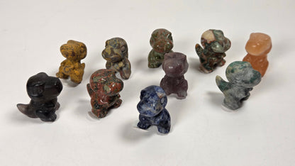 A group of small carved stone figurines shaped like dinosaurs, displayed on a plain white surface. The figurines are made from various natural stones in different colors, including black, brown, green, blue, purple, and orange.