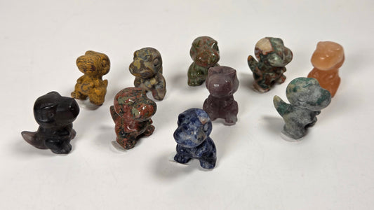 A group of small carved stone figurines shaped like dinosaurs, displayed on a plain white surface. The figurines are made from various natural stones in different colors, including black, brown, green, blue, purple, and orange.
