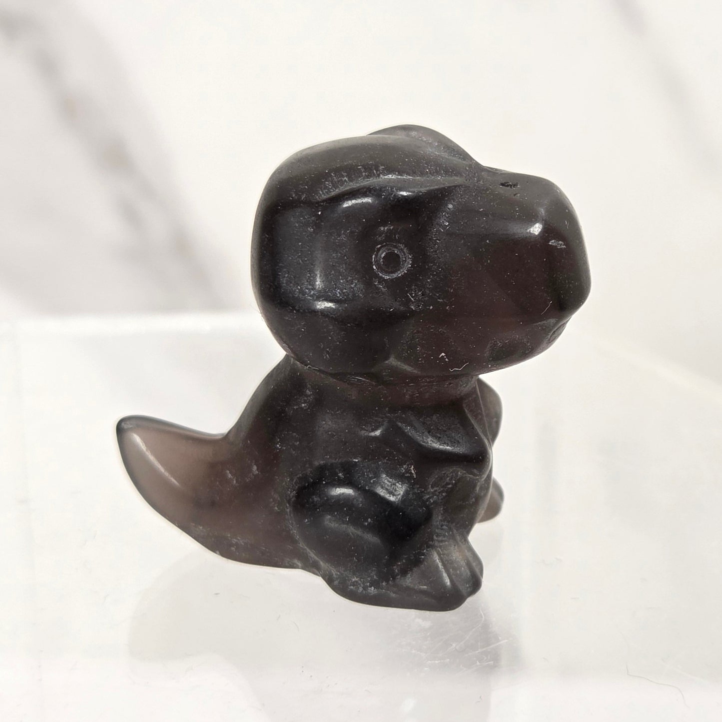 A small carved dark brown or near-black figurine shaped like a seated dinosaur or creature, displayed on a clear acrylic stand against a light marble-like background.