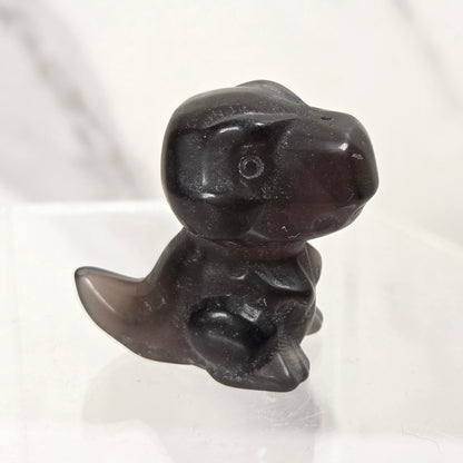 A small carved dark brown or near-black figurine shaped like a seated dinosaur or creature, displayed on a clear acrylic stand against a light marble-like background.