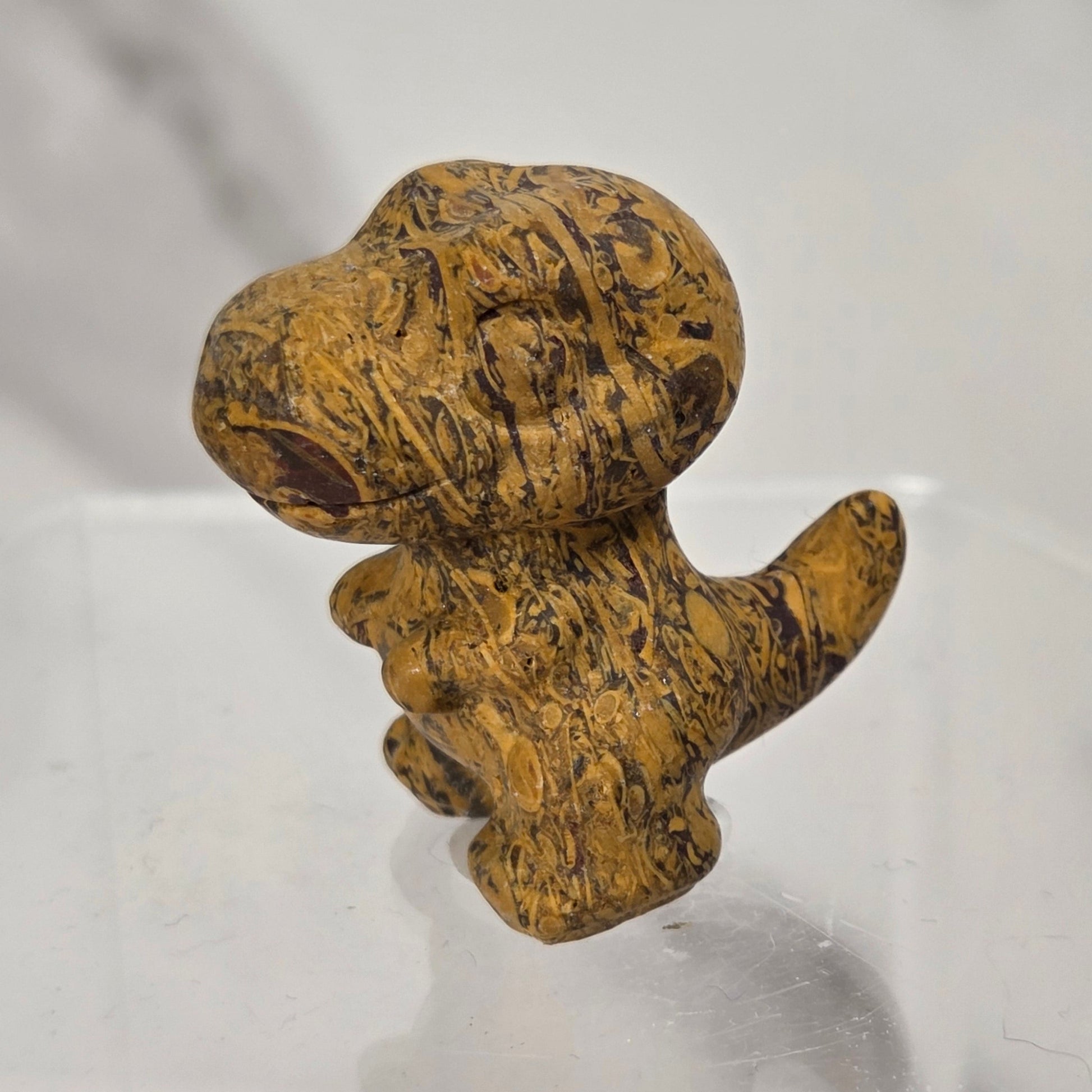 A small carved figurine of a dog made from textured golden-brown wood with dark grain patterns, standing on a clear acrylic display block against a light gray marble-patterned background.