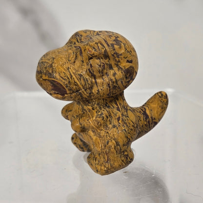 A small carved figurine of a dog made from textured golden-brown wood with dark grain patterns, standing on a clear acrylic display block against a light gray marble-patterned background.
