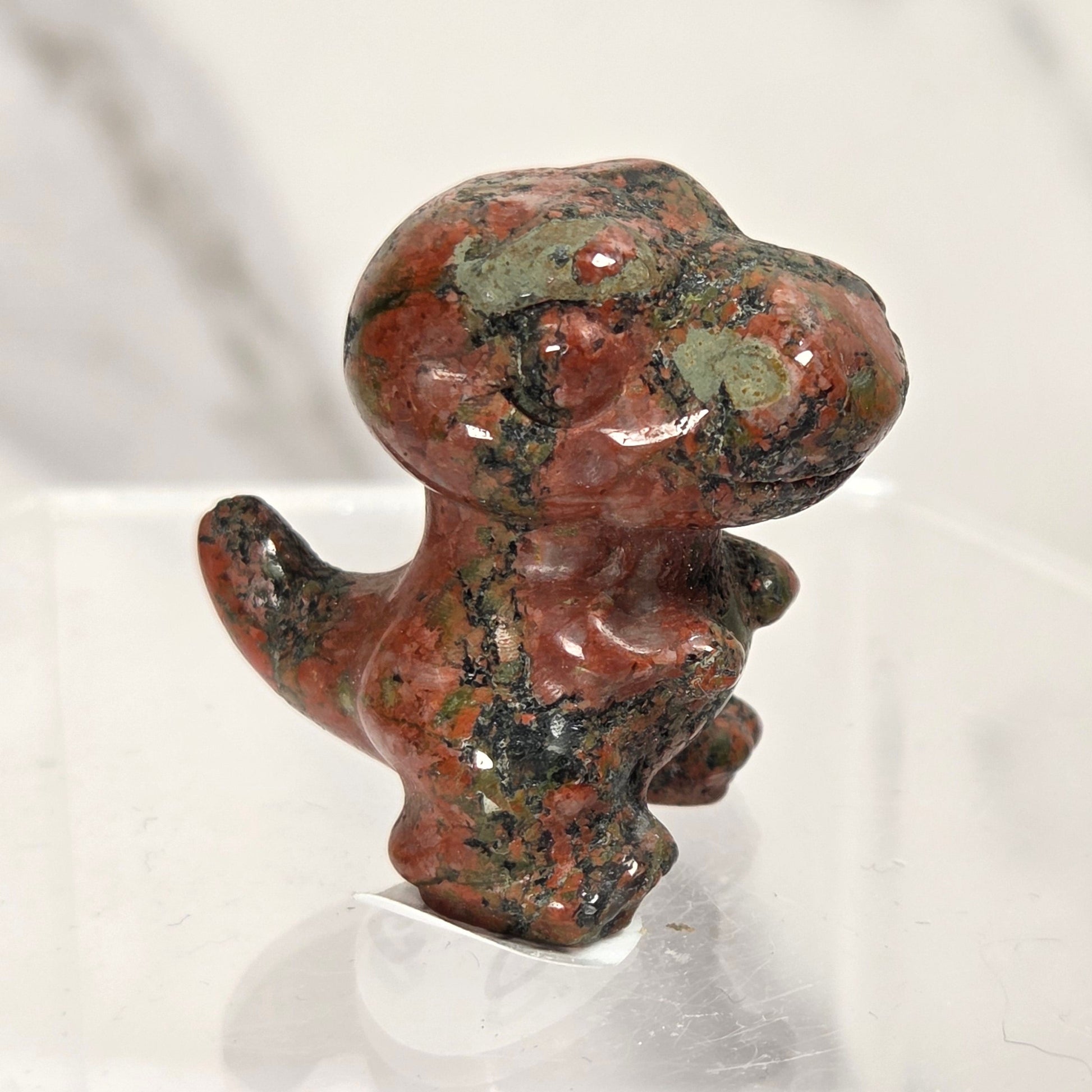 A small polished stone figurine of a cartoon character, likely a dinosaur or creature, featuring a reddish-brown and green mottled pattern, displayed on a clear acrylic stand.