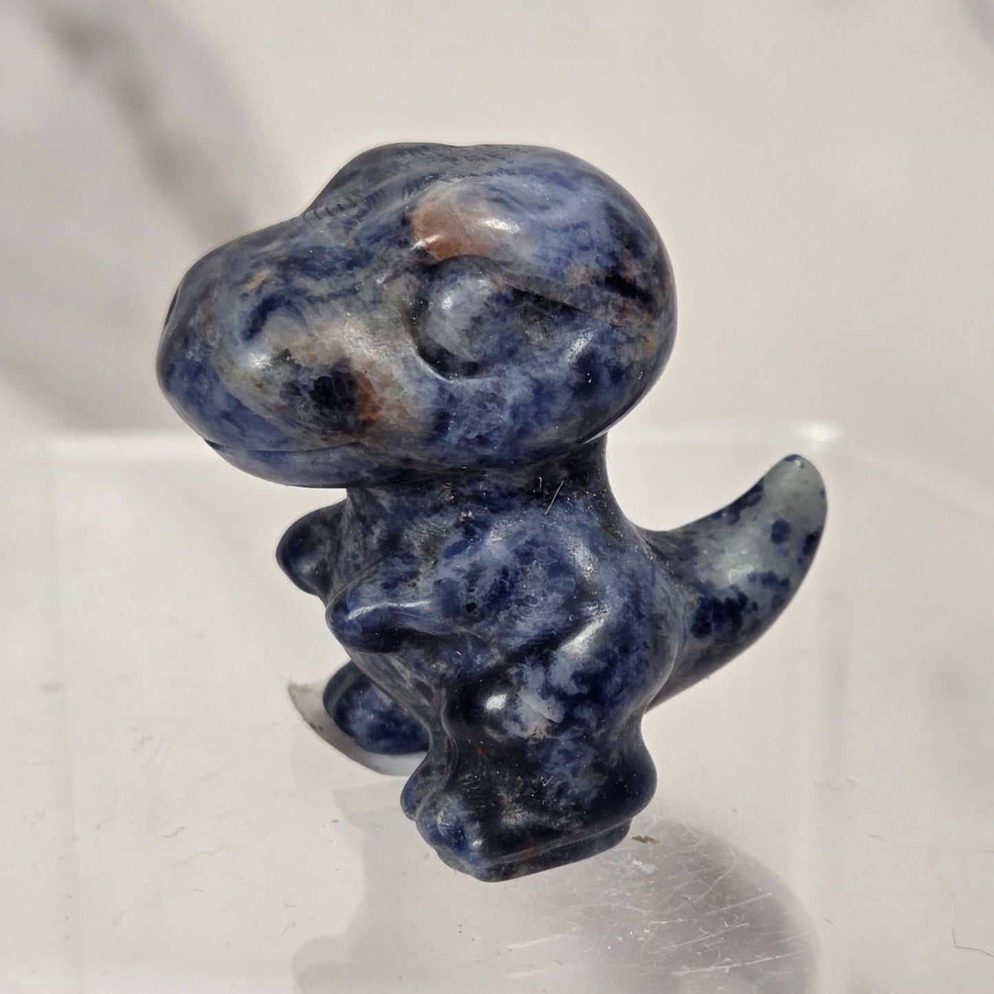 A small carved figurine of a dinosaur or dragon made from polished blue stone with white and dark marbling, standing on a clear acrylic display block against a light marble-like background.