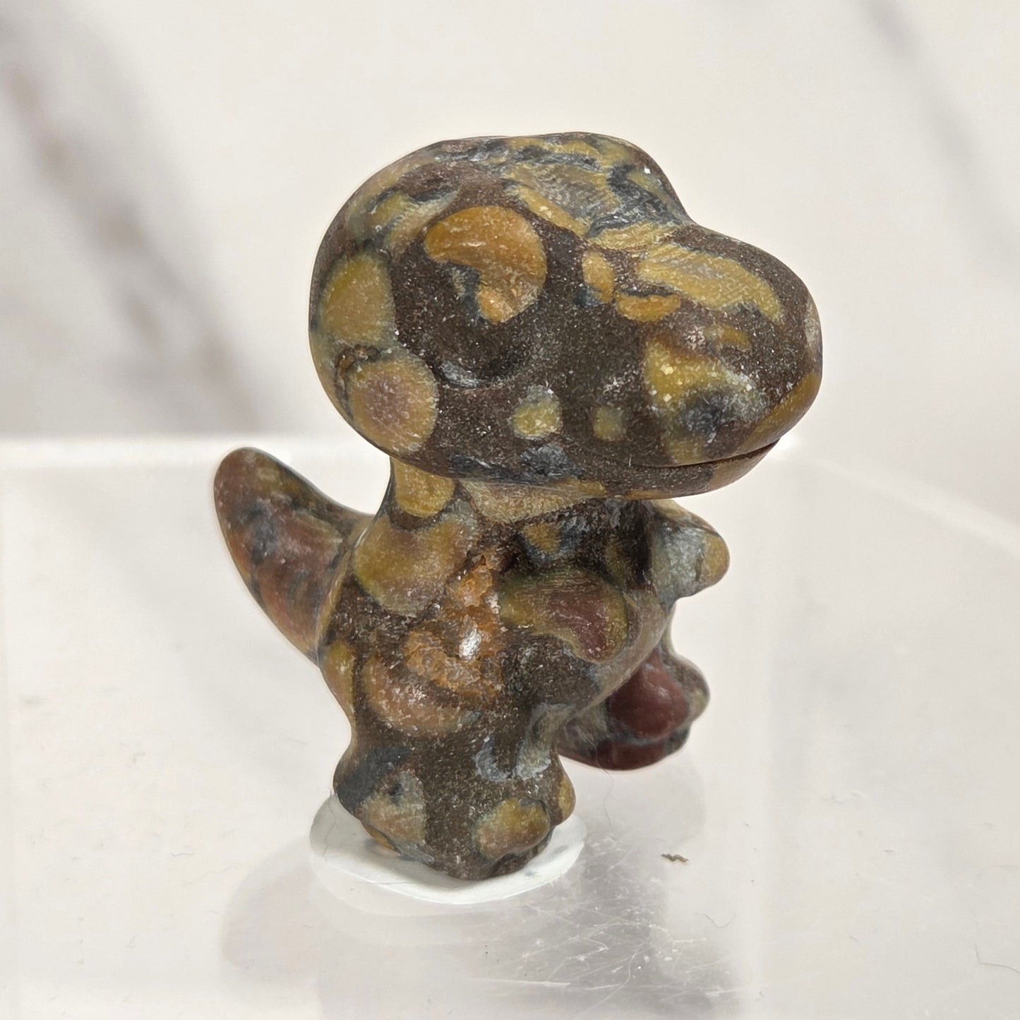 A small sculpted figurine of a dinosaur or reptile-like creature with a rounded head and short legs, made from polished stone with mottled brown, tan, and dark gray patterns, displayed on a clear acrylic riser against a light marble-patterned background.