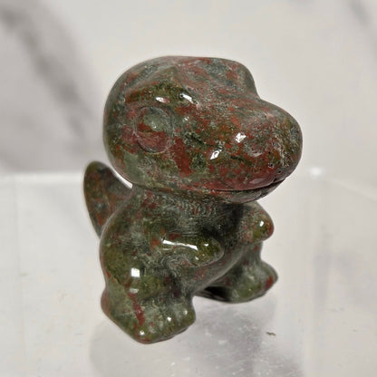 A small polished stone figurine shaped like a dinosaur, featuring a mottled pattern of green, red, and gray, displayed on a clear acrylic stand against a light gray marble-patterned background.