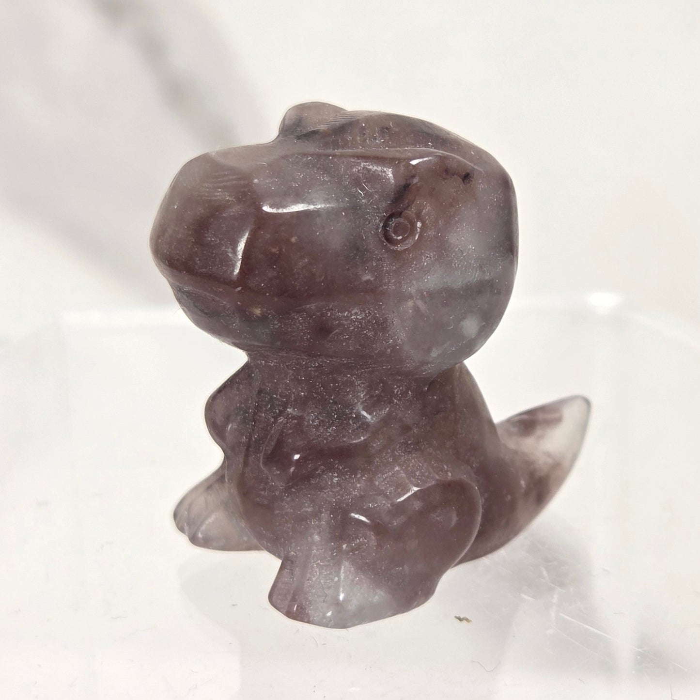 A small carved stone figurine of a dinosaur or reptile character, made from semi-translucent dark gray and purple stone, displayed on a clear acrylic stand against a light marble-patterned background.