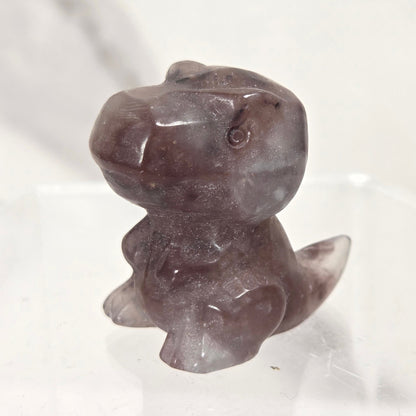 A small carved stone figurine of a dinosaur or reptile character, made from semi-translucent dark gray and purple stone, displayed on a clear acrylic stand against a light marble-patterned background.