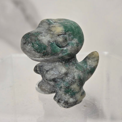 A small sculpted dinosaur figurine made from marbled green and white stone, standing on a clear acrylic display base against a light marble-patterned background.