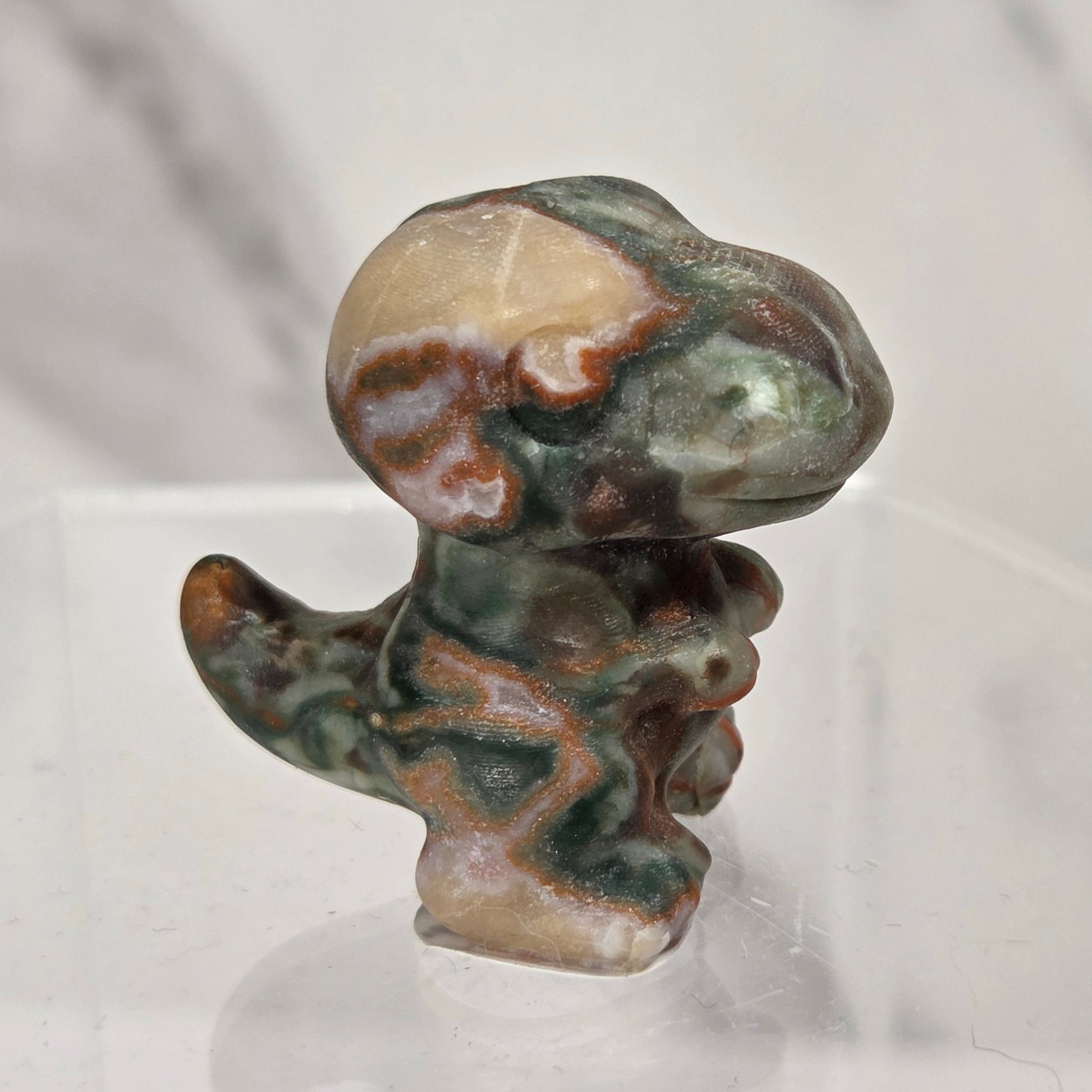 A small sculpted figurine of a dinosaur made from polished marbled stone, featuring green, brown, beige, and gray tones, displayed on a clear acrylic stand against a light gray marble-patterned background.